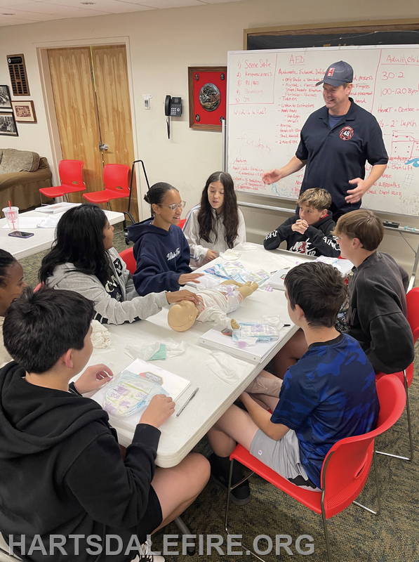 A group of students learns about first aid, practicing on a dummy, while an instructor teaches in a classroom setting.