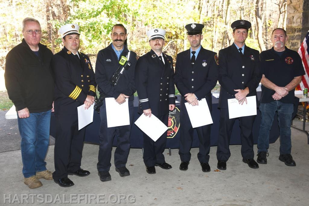 A group of uniformed firefighters and officials stand together at a ceremony, holding documents, with a scenic background.