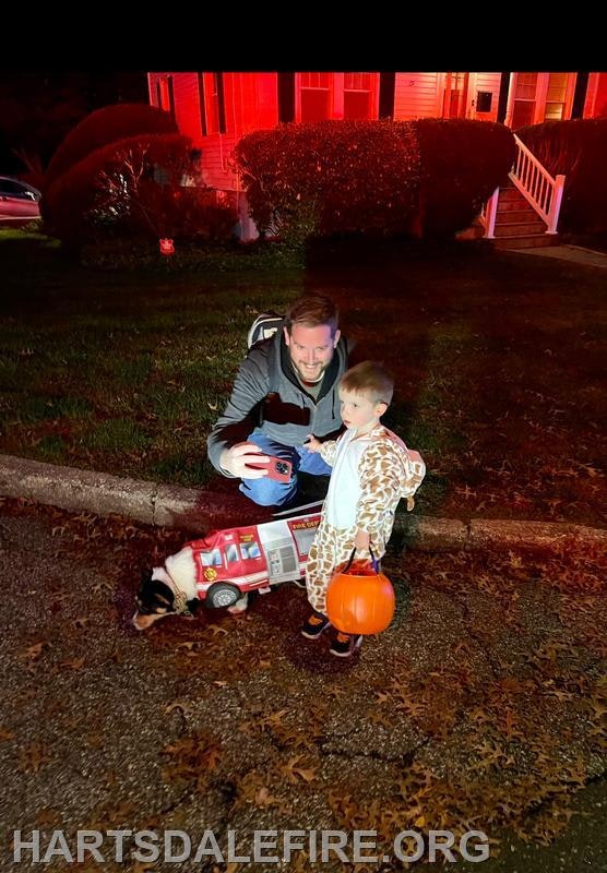 A man and a child in a costume pose for a photo at night, with a toy fire truck and a dog nearby.