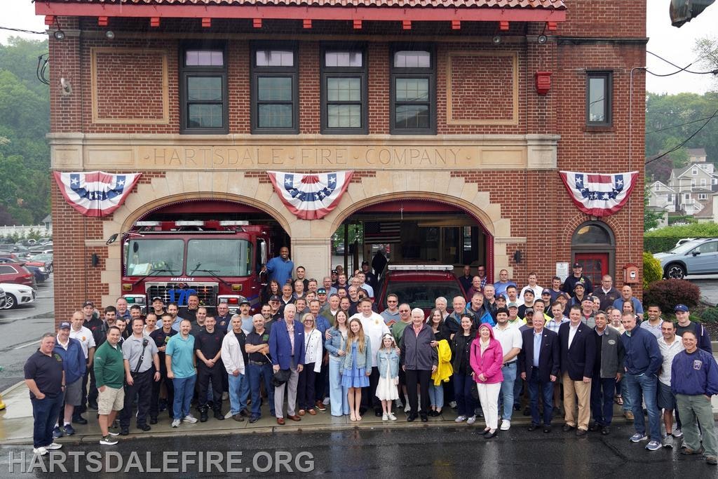The image shows a large group of people gathered outside the Hartsdale Fire Company with fire trucks in the background.