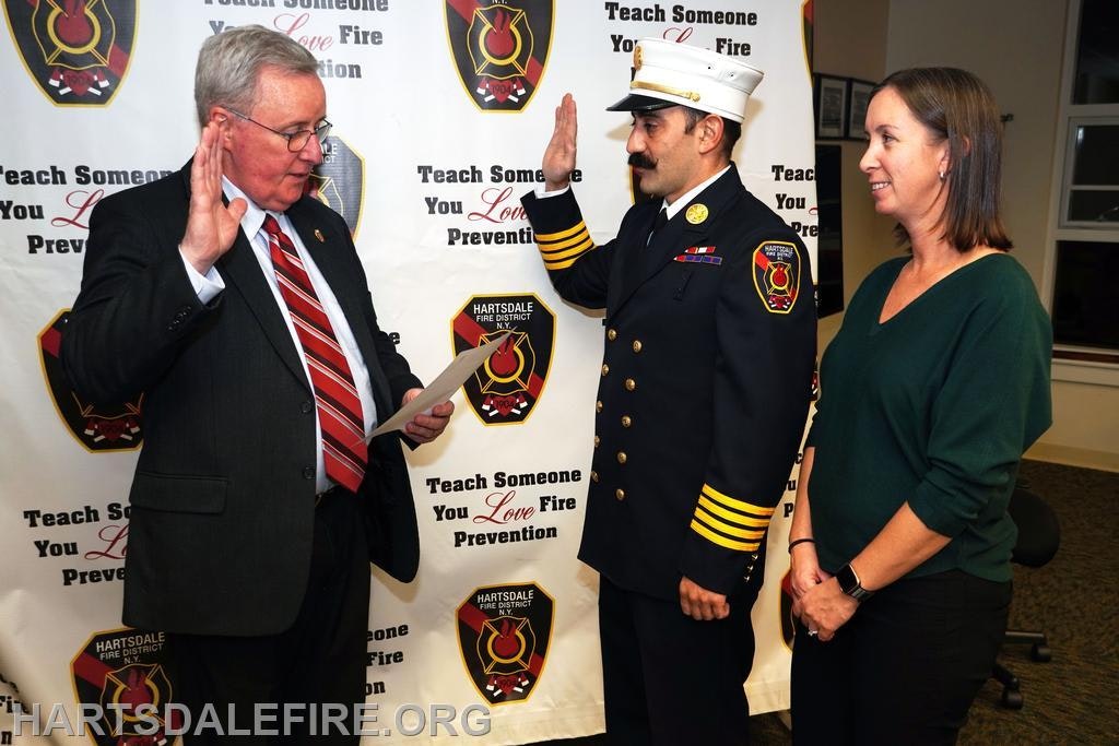 A fire chief is being sworn in at a ceremony, supported by a woman and a man reading from a document.