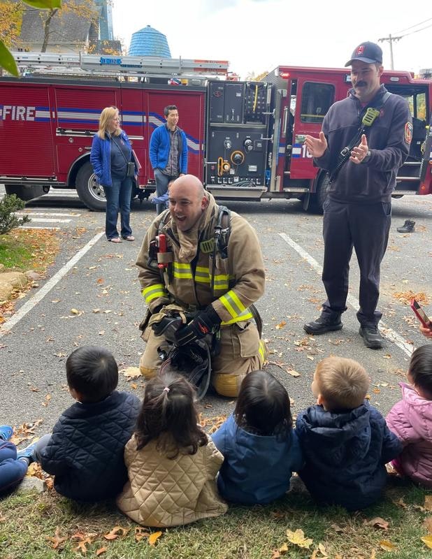 A firefighter engages with young kids while another firefighter and adults observe nearby, with a fire truck in the background.