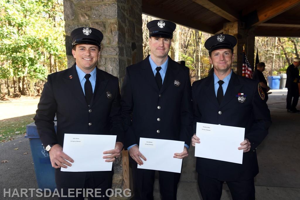 Three uniformed firefighters stand together holding blank certificates outdoors, with trees and an event setup in the background.
