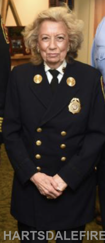 The image shows a woman in a formal fire department uniform with insignia, standing with her hands clasped.