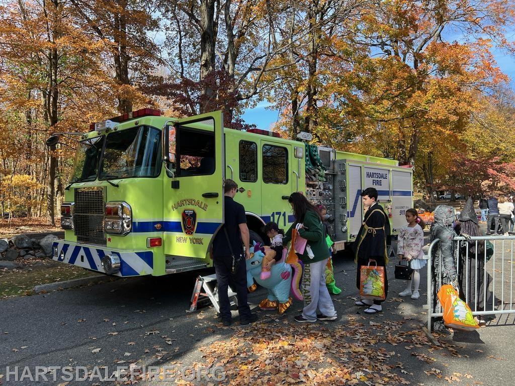 A vibrant scene with a green fire truck in a fall setting, featuring children in costumes and adults engaging in community activities.
