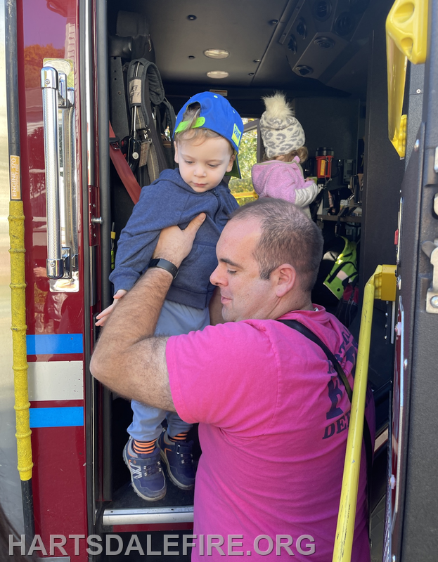 A firefighter assists a young child stepping onto a fire truck, with another child visible in the background.