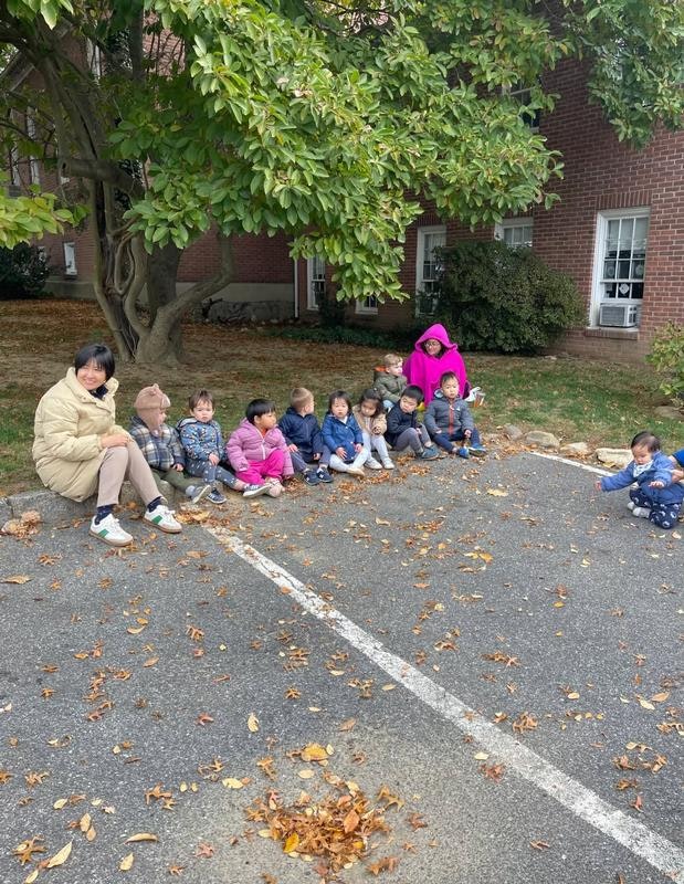 A group of children sitting on a sidewalk under a tree, with two adults nearby, surrounded by fall leaves.