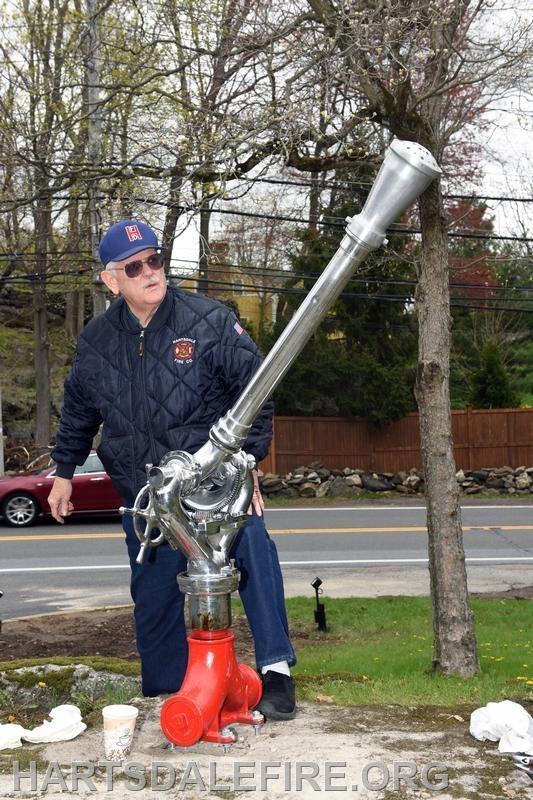 An elderly man in a fire department jacket stands beside a shiny fire hydrant, adjusting its aiming nozzle outdoors.