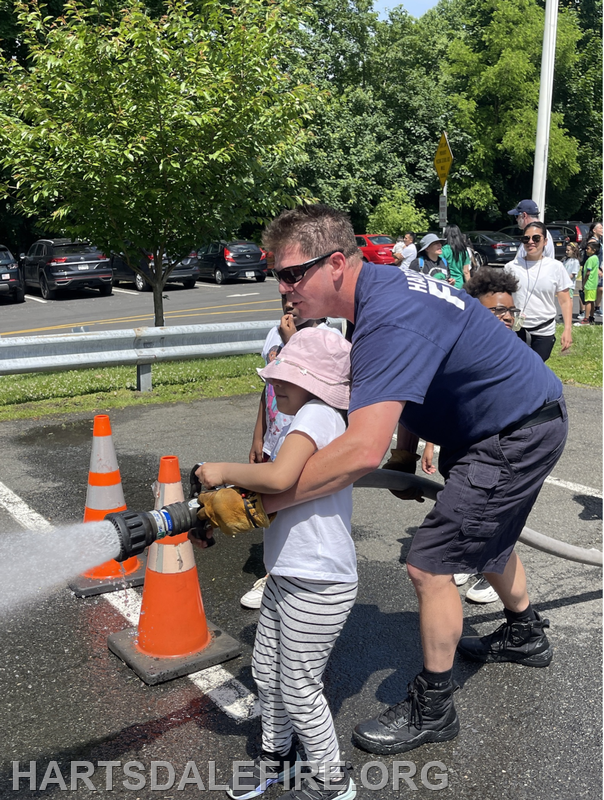 A firefighter guides a child using a fire hose, demonstrating safety and skills at an event, with onlookers nearby.