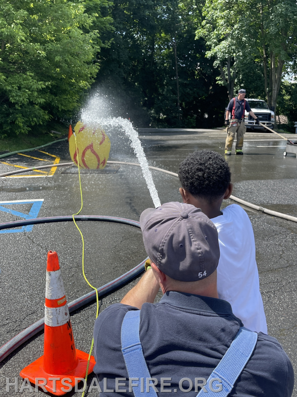 A child learns to use a fire hose, spraying water at a target while a firefighter supervises in a training scene.
