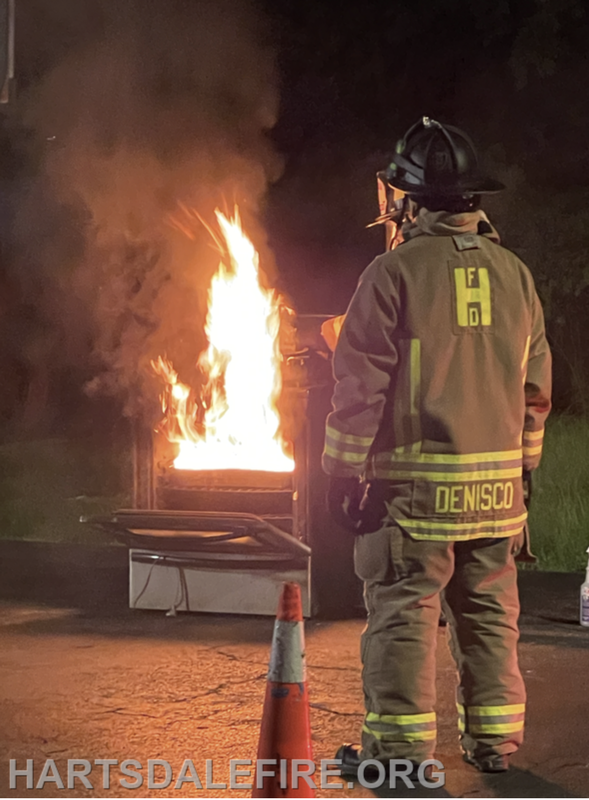 A firefighter in gear stands near a burning dumpster at night, observing the flames and smoke. Safety cone nearby.