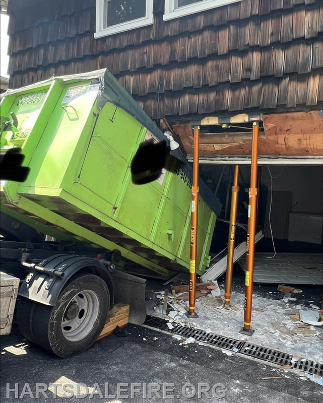 A green dumpster is tipped against a damaged house, with supports holding up the structure. Debris is scattered around.