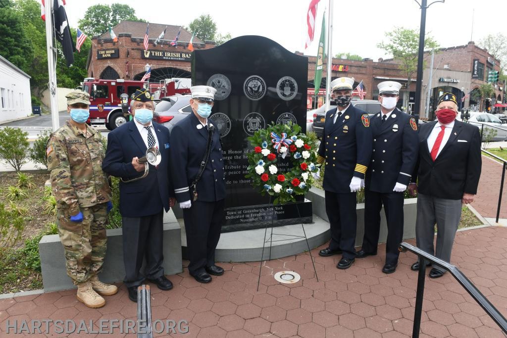 A group of people in uniforms stands by a memorial with a wreath. They are wearing masks, and a fire truck is visible in the background.