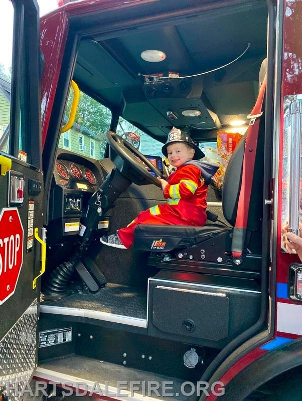 A child in a firefighter costume sits in the driver's seat of a fire truck, smiling and enjoying the moment.