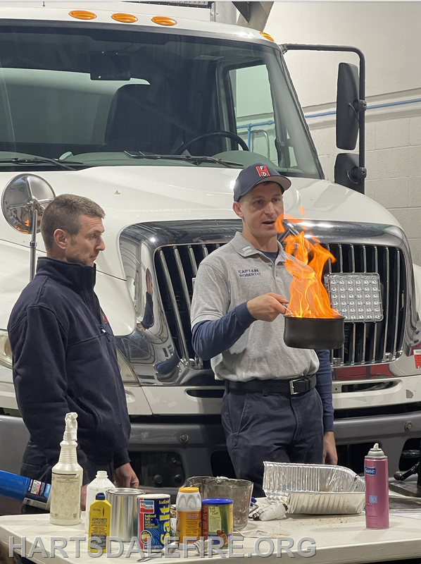 Two men are demonstrating fire safety, one holding a flaming pot in front of a truck, while various materials are on the table.