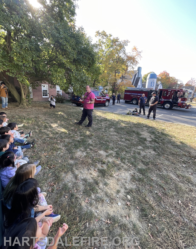 A group of children sits on the grass, watching a fire safety demonstration by a speaker near fire trucks and emergency personnel.
