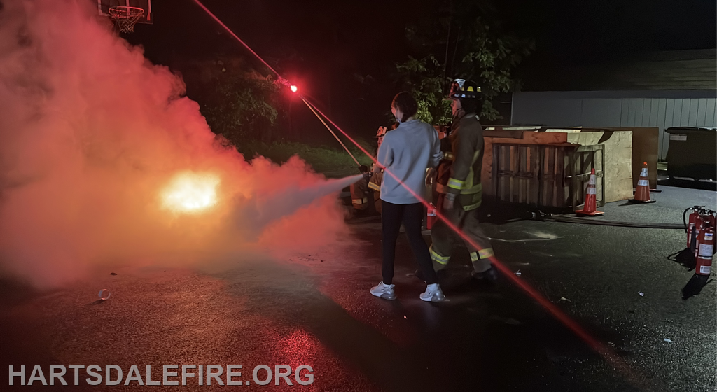 A firefighter demonstrates fire control techniques at night, surrounded by smoke and equipment, with a bystander observing.