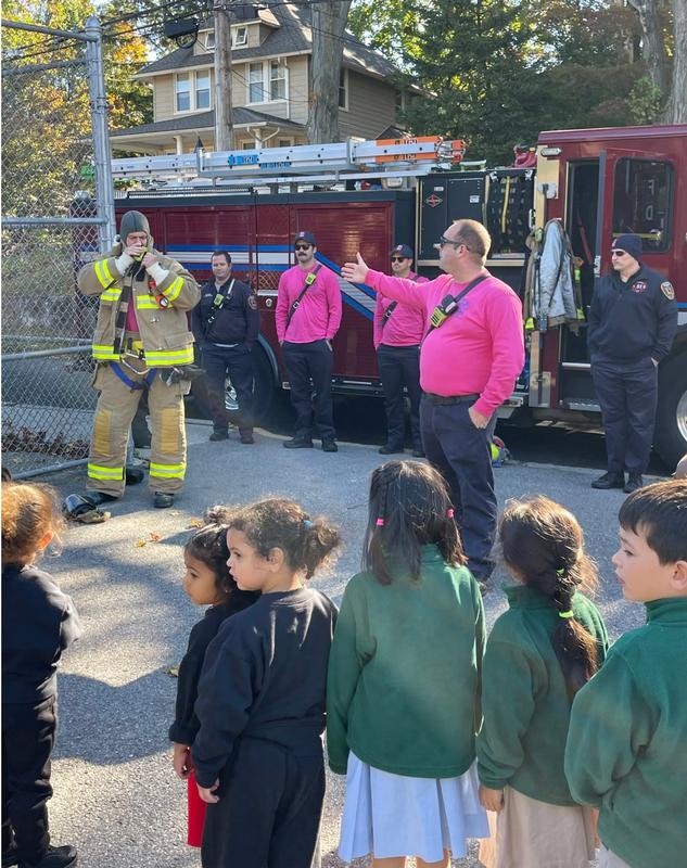 A group of children listen to firefighters in pink shirts near a fire truck, promoting fire safety and community engagement.