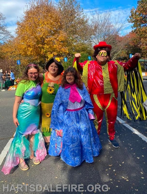 A group of four people in colorful costumes posing outdoors in autumn. Trees with orange leaves are in the background.