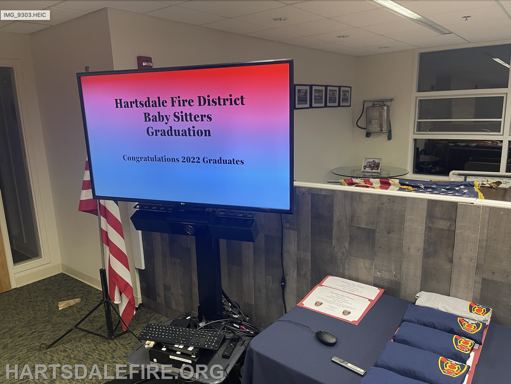 TV displays "Hartsdale Fire District Baby Sitters Graduation." Table with certificates and shirts nearby.