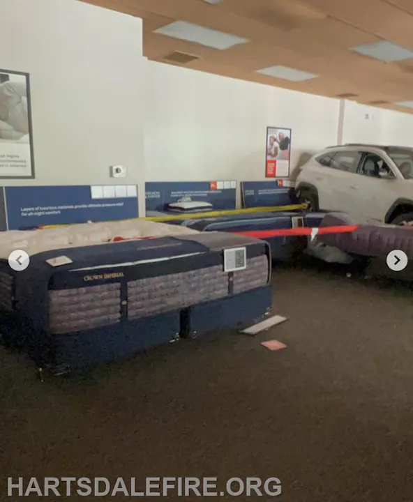 The image shows a mattress showroom with several beds displayed and an SUV partially parked among them.