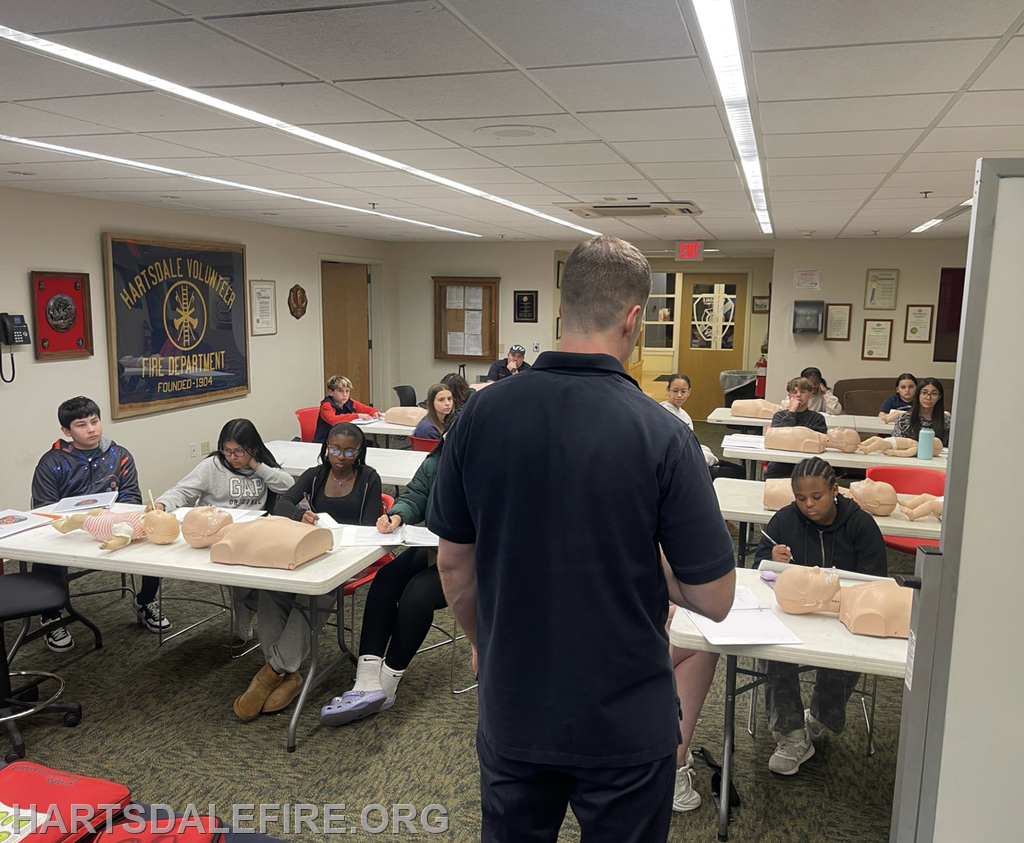 A group of students is in a classroom learning about CPR, with training mannequins on the tables and an instructor leading the session.