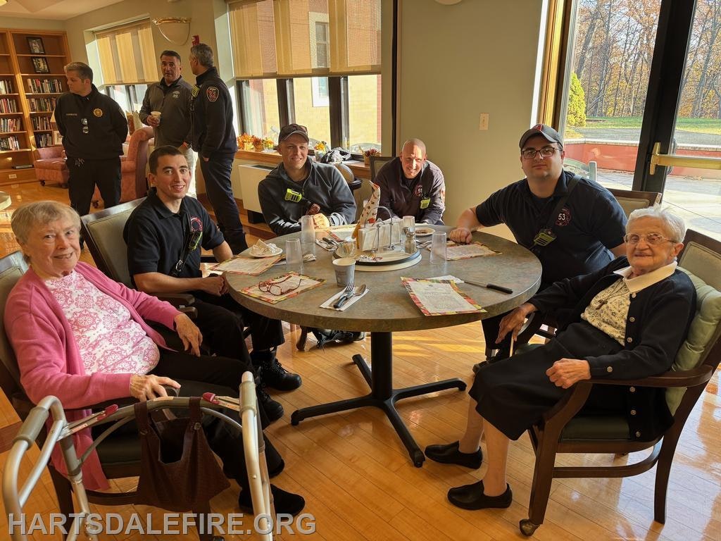 A group of elderly women and firefighters gather around a table, enjoying a friendly interaction in a cozy setting.