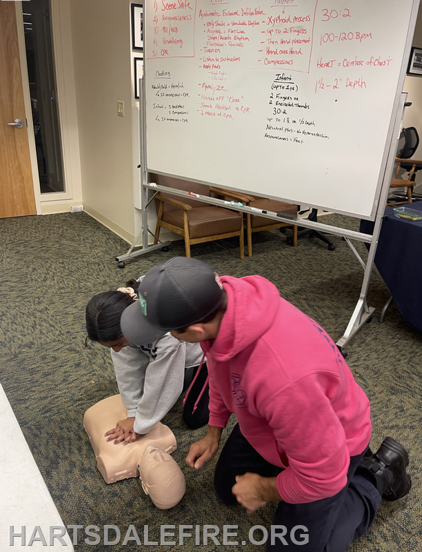 A training session on CPR, with two individuals practicing on a manikin, while notes are visible on a whiteboard.
