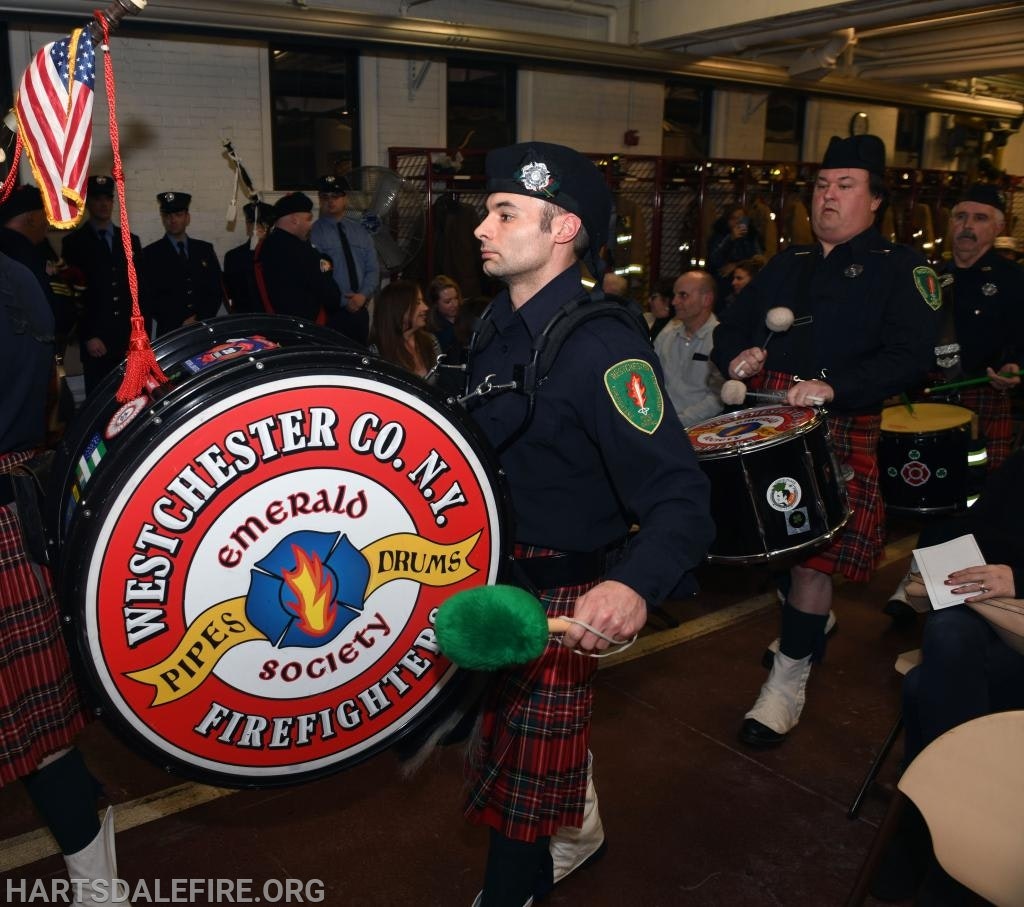 A firefighter pipe band in uniforms and kilts playing drums, with an American flag and audience in the background.