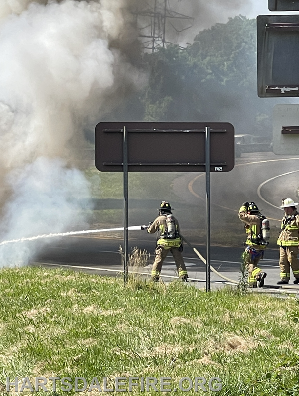 Firefighters are battling a fire alongside a road, with smoke billowing into the air.