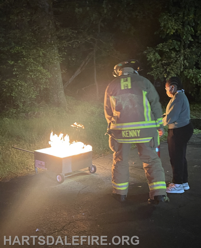 A firefighter in gear attends to a woman standing near a burning cart in a dark outside setting.