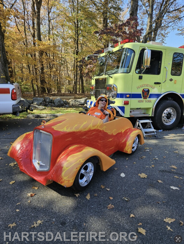 A person in an orange outfit enjoys a fun, flame-decorated toy car near a fire truck in a forested area.