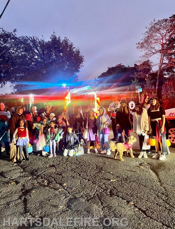 A group of children and adults in costumes poses in front of a fire truck, illuminated by its lights during a nighttime event.