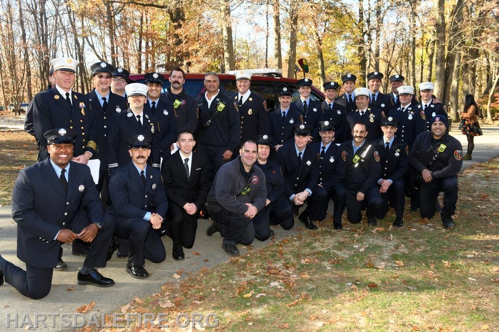 A group of firefighters in uniform poses together outdoors, surrounded by autumn trees.