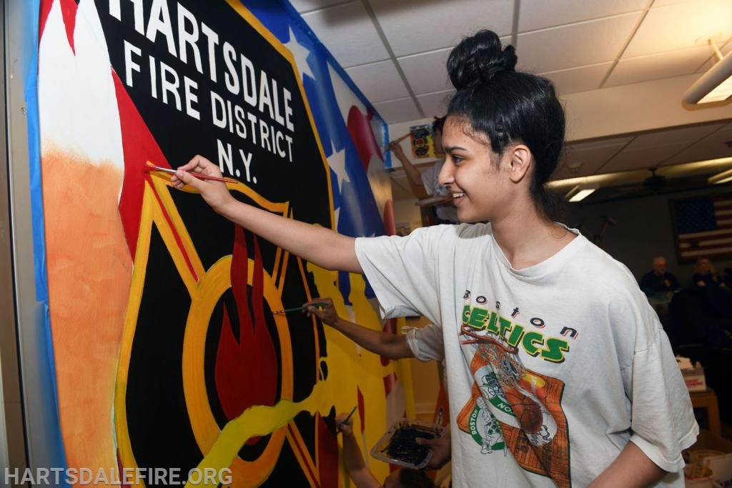 A person is painting a mural featuring "Hartsdale Fire District N.Y." The scene is indoors with an American flag in the background.