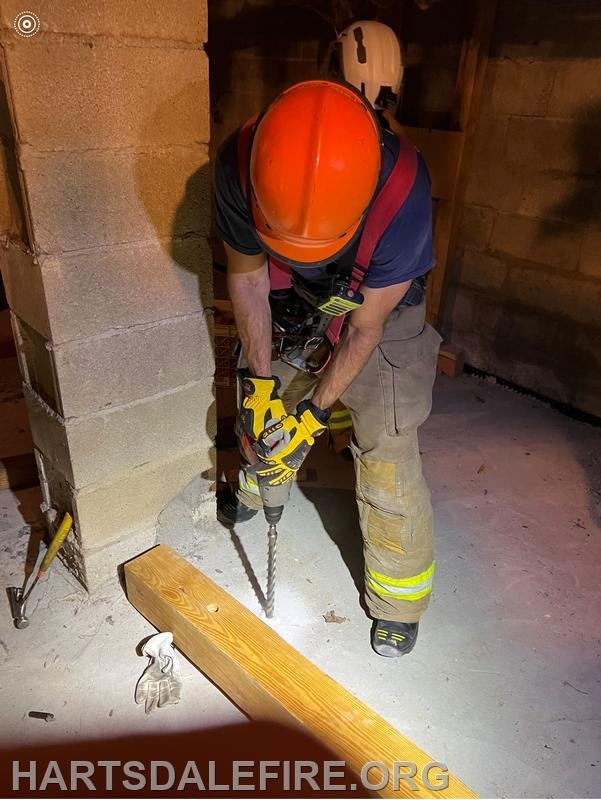A person in safety gear is using a power drill on a wooden beam in a construction or training setting.