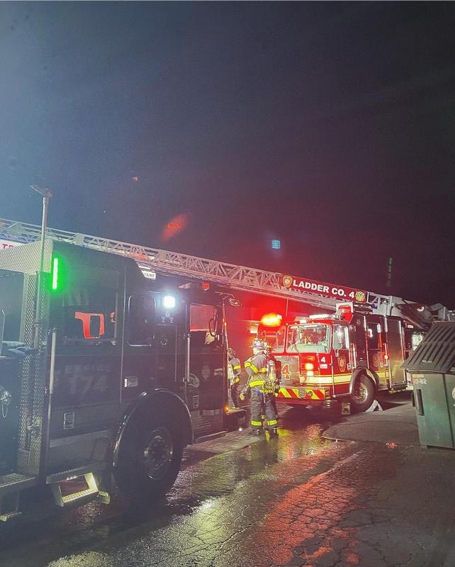 A fire scene at night with fire trucks and a firefighter near a ladder truck, illuminated by emergency lights.
