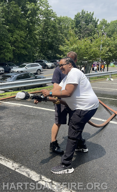 Two people are operating a fire hose in a parking lot, enjoying the activity amidst a green backdrop.
