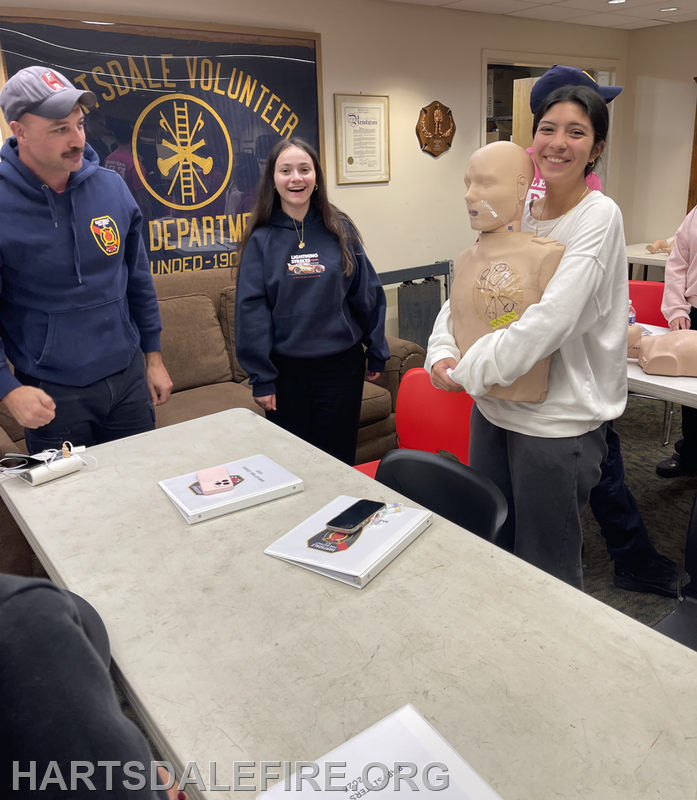 The image shows a training session at Hartsdale Volunteer Fire Department, with participants engaging with a practice mannequin.