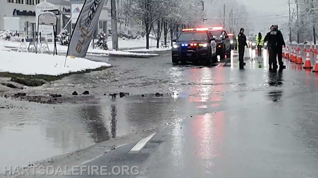 The image shows a snowy road with police cars, workers, and caution cones near a water leak or flooding situation.