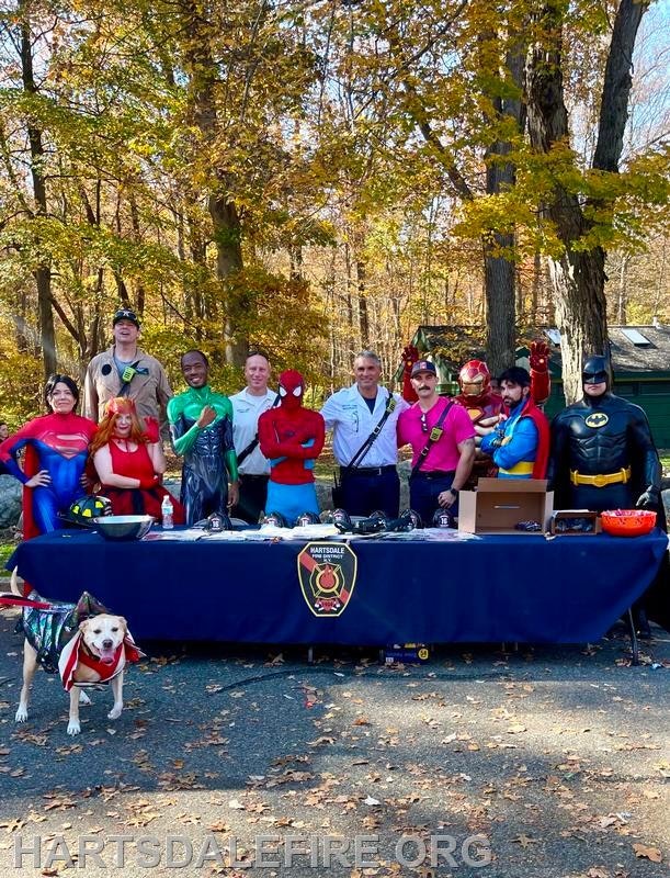 A group of people in superhero costumes poses by a table, with a dog, in a colorful autumn setting.
