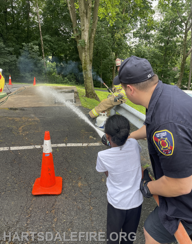 A firefighter demonstrates equipment usage to a child, with water spraying and safety cones marking the area.
