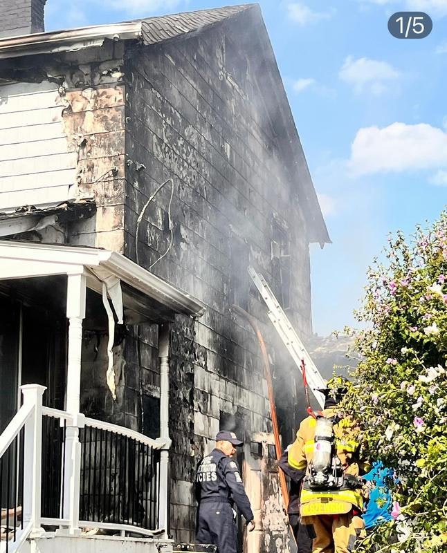 Firefighters are responding to a serious fire on a house, with smoke billowing and visible damage to the structure.