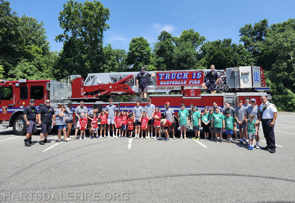 A group of children and firefighters pose together in front of a fire truck on a sunny day.