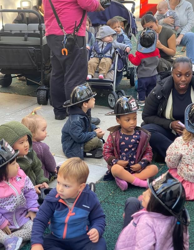 A group of young children wearing firefighter helmets are sitting on the ground, enjoying a playful gathering.