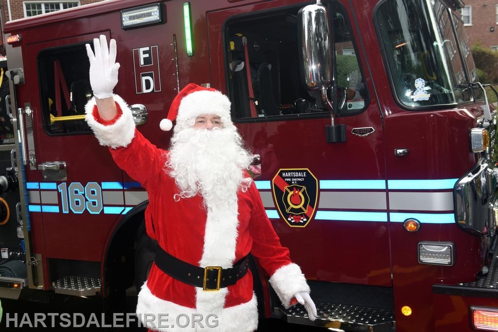 A person dressed as Santa Claus waves in front of a fire truck with a fire department logo.