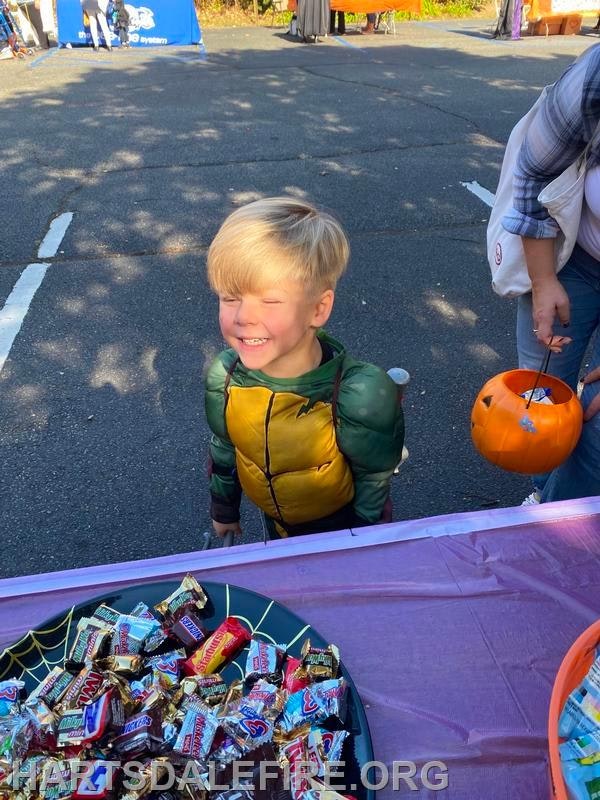 A smiling child in a costume stands near a table filled with candy at an outdoor event, holding a pumpkin-shaped candy bucket.