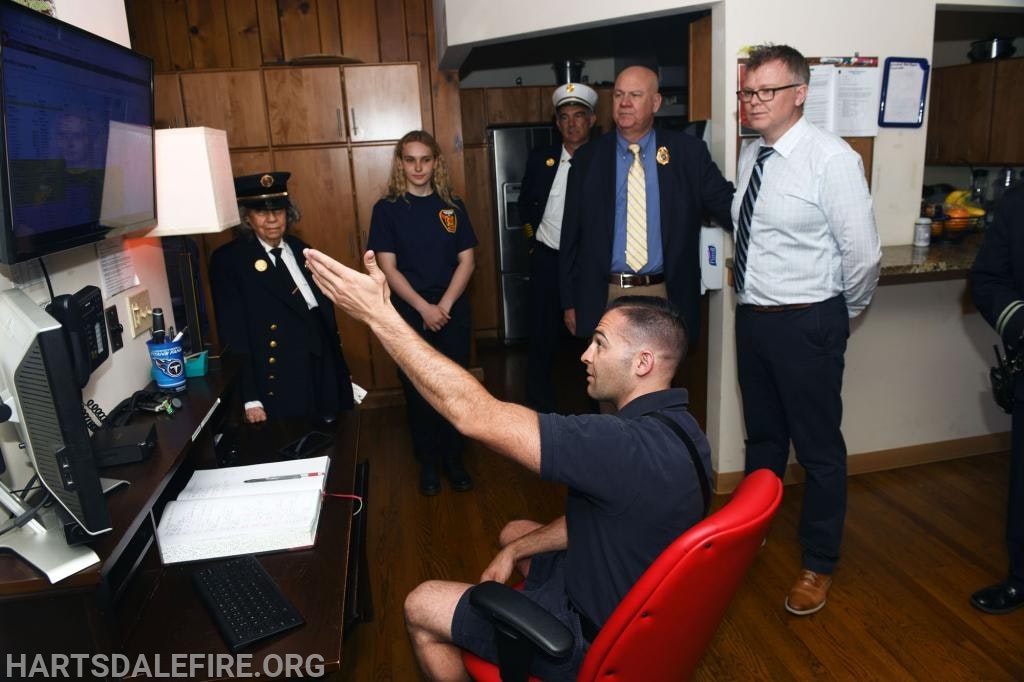 A group of people, including a person in a fire uniform seated at a computer, are in an office setting looking at a screen.