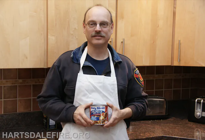 A man wearing an apron holds a can of Dinty Moore beef stew in a kitchen setting.