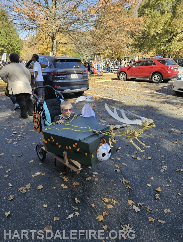 A child in sunglasses rides a decorated wagon featuring Halloween themes, including skeletons and pumpkins, amidst a festive crowd.
